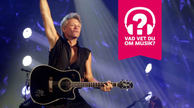 Jon Bon Jovi har en akustisk gitarr kring halsen och han sträcker höger arm rakt upp i luften.