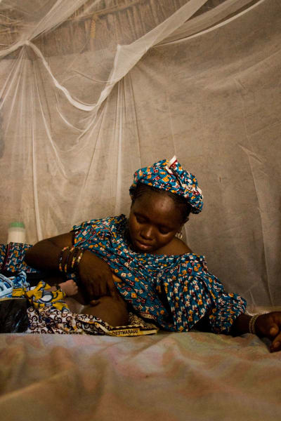 En kvinna ammar sitt barn under ett myggnät i Afrika. De ligger på golvet.