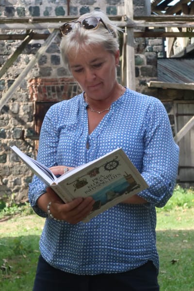 Carina Wolff-Brandt står bland gamla slaggstensbyggnader med boken uppslagen i händerna och läser ur boken.