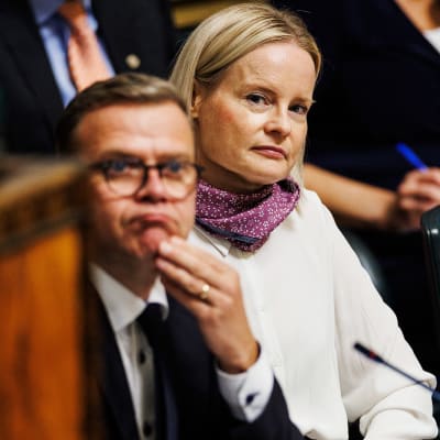 Petteri Orpo ser fundersam ut i ministerbåset i riksdagen, Riikka Purra sneglar fram över hans axel.