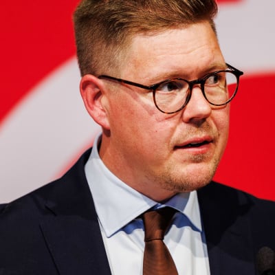 En man med runda glasögon, iklädd en mörkblå kostym, ser bekymrad ut. Han står på en scen, och bakgrunden är röd och vit.