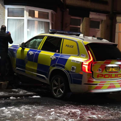 En brittisk polisbil har kört in i ett bostadshus. En man granskar skadorna på huset med hjälp av en ficklampa. Marken bakom bilen är frusen.