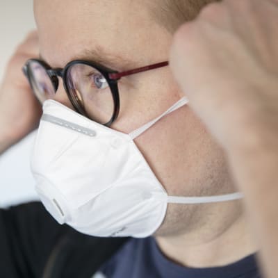 En person justerar sitt andningsskydd vid öronen. Personen har glasögon och en svart tröja på sig och hens ögon är blå.