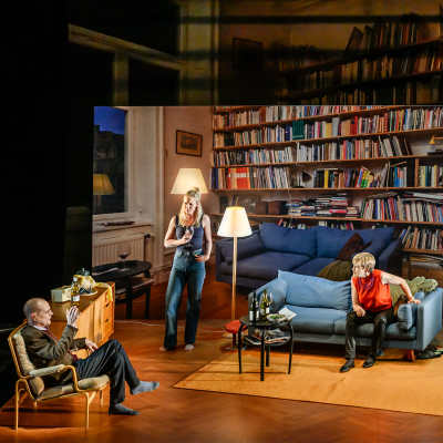 Tre skådespelare sitter i en soffgrupp på en scen inredd som ett vardagsrum med en stor bokhylla i bakgrunden. 