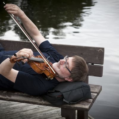 Mies makaa selällään penkillä järven rannassa ja soittee viulua sylissään.