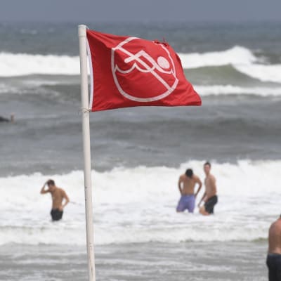 I förgrunden en röd flagga som signalerar att man inte ska simma, i bakgrunden en strand och folk som simmar i vattnet.