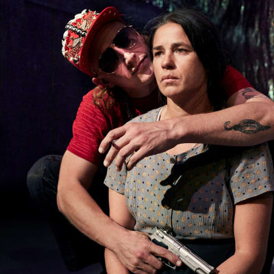 En man i solglasögon och pistol håller om en ung kvinna. 