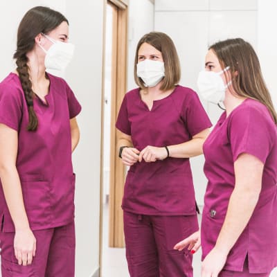 Tre kvinnliga sjukskötare i munskydd och uniform står och pratar med varandra i en korridor.