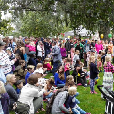 Många barn och vuxna står i en park och lyssnar på en konsert.