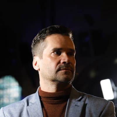 Mika Aaltola puolilähikuvassa tummaa taustaa vasten, katsoo omalle vasemmalle puolelleen.