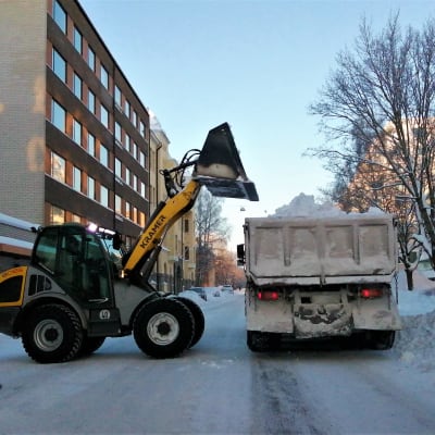 Snöplog lyfter upp lass med snö på flaket till en lastbil längs Skolhusgatan i Vasa.