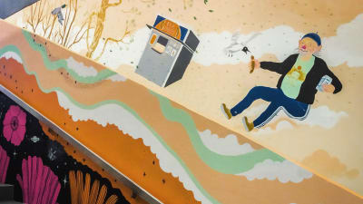 Seinämaalaus Martinlaakson juna-aseman sisäseinällä, mies jolla kädessä nakki, lentävä lokki, pesukone.