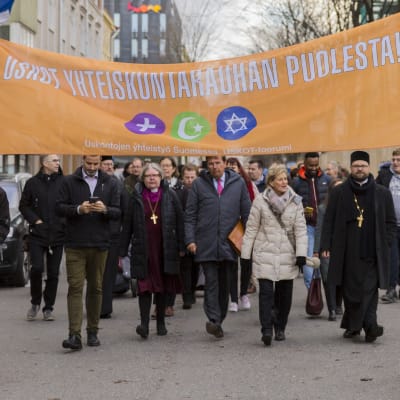 Eri uskontokuntiin kuuluvien yhteinen rauhankävely. Banderollissa teksti uskot yhteiskuntarauhan puolesta.