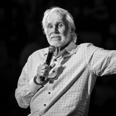 Kenny Rogers står på scen med en mikrofon i ena handen och andra armen utsträckt. Bilden är tagen i Austin, Texas under hans “Farewell Tour” den 19 mars 2017.