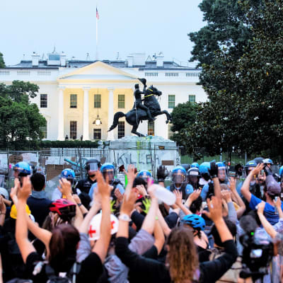 Mielenosoittajien joukko seisoo mellakkavarusteissa olevan poliisiketjun edessä ja pitää käsiään ylhäällä. Taustalla näkyy Valkoinen talo ja Andrew Jacksonin patsas.