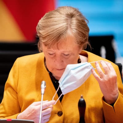 Angela Merkel, klädd i gult, tar av sig sitt munskydd framför en mikrofon.