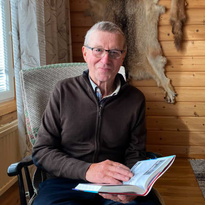 Voitto Kovakka istuu keinutuolissa kirja kädessään ja katsoo kameraan.