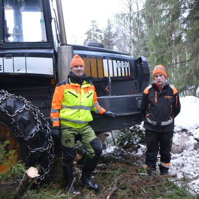 Två män står framför en traktor. De befinner sig i en snöbeklädd skog. Den ena av männen har en gul rock på sig och båda har arbetskläder.