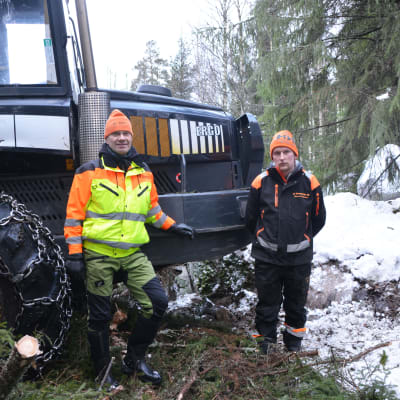 Två män står framför en traktor. De befinner sig i en snöbeklädd skog. Den ena av männen har en gul rock på sig och båda har arbetskläder.