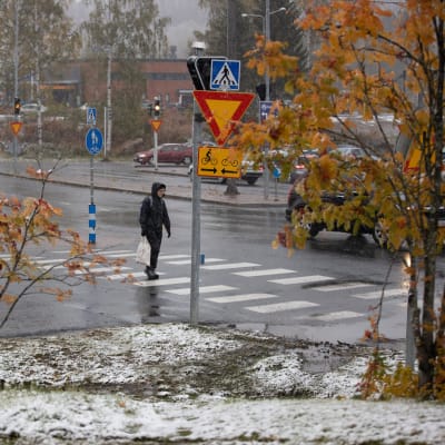 En människa klätt i mörka kläder går över en gatukorsning. Det är regnvått på gatan, men på gräsmattorna runt omkring ligger ett lätt snötäcke och det är fortfarande löv på träden omkring.