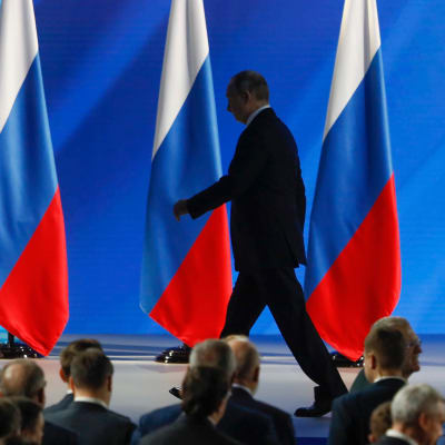 I bakgrunden flera ryska flaggor, framför den en kostymklädd man som går och i förgrunden huvuden tillhörande en publik.  Rysslands president Vladimir Putin lämnar podiet efter sitt tal till nationen den 15 januari 2020.