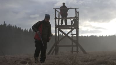 Joonas Saartamo med gevär i handen går mot kameran medan Peter Franzén håller utkik från ett jakttorn.