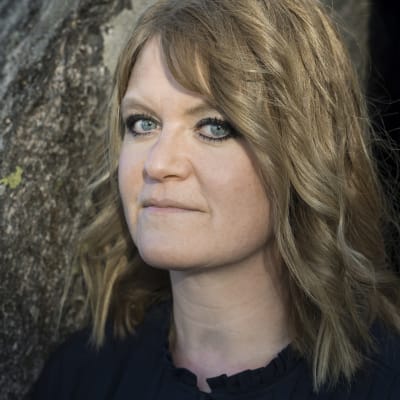 En kvinna med svart eyeliner står framför en trädstam och ser in i kameran. 