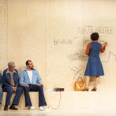 En kvinna står på en bänk och skriver på väggen med tusch. Tre personer på sidan av och betraktar henne. 