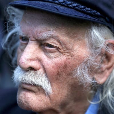 Valkohiuksinen viiksekäs Manolis Glezos katsoo kuvassa tuimasti kuvaajasta vasemmalle.
