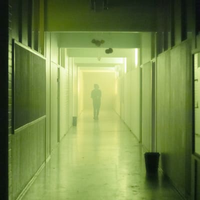 En disig och dunkelt upplyst skolkorridor. Längst borta i korridoren syns siluetten av en gående person. 