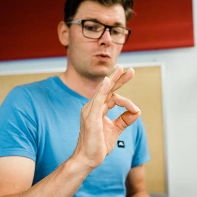 En man i blå t-shirt visar teckenspråkstecknet för fråga.