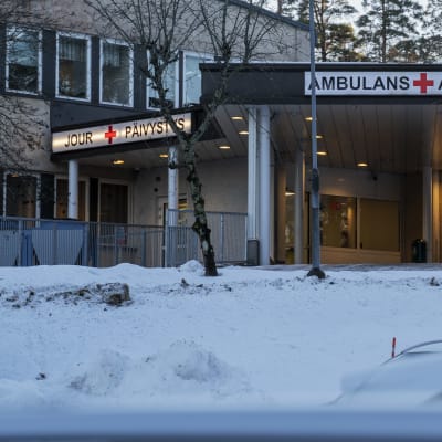 En sjukhusbyggnad med en stor skylt där det står ambulans på svenska och finska. I förgrunden syns taket av några bilar.