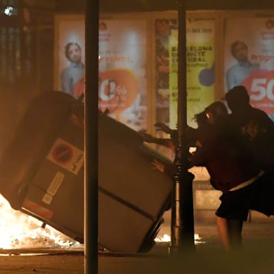 Demonstranter fäller en soptunna i Barcelona. Eld i bakgrunden.