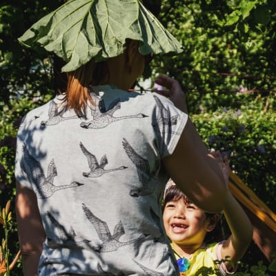 Liten flicka ler och sträcker upp armarna mot en vuxen som står med ryggen mot tittaren, och håller ett rabarberblad som hatt på huvudet.