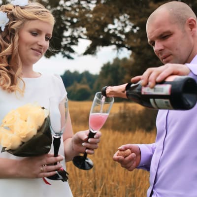 Ung man och kvinna i bröllopskläder står på äng och häller upp champagne i glas
