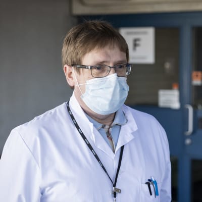 Mansperson i läkarrock och ansiktsmask står utomhus vid sjukhusentré.