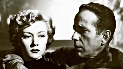 Ilmeettömän näköinen nainen (Gloria Grahame) istuu auton etupenkillä, mies (Humphrey Bogart) katsoo häntä vierestä ja pitää kättä naisen olalla. Kuva elokuvasta Hermot pinnalla (In a Lonely Place).
