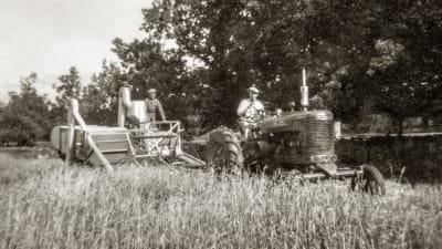 ett gammalt svartvitt fotografi. Två män, en på traktor, andra på en gammeltida skördetröska på ett sädesfält. Sensommar..