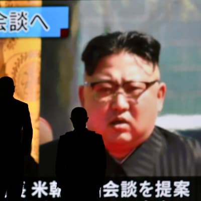 Ihmiset kävelevät suuren televisionäytön ohi. Näytöllä näkyy arkistomateriaalia Donald Trumpista ja Kim Jong-unista.