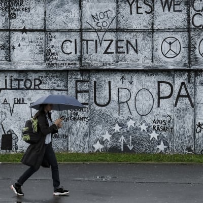 En mur med väggmålningar med EU-tema i rumänska staden Sibiu. I förgrunden en förbipasserande  invånare med ett paraply i handen. 