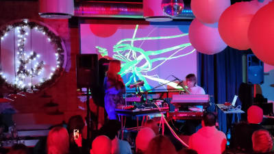 Konsert, tre musiker, synthar, ballonger i taket. publik. 