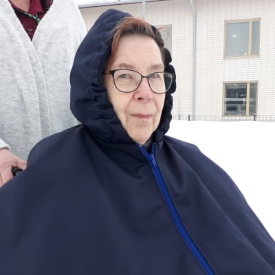 Mirja Malmi istuu pyörätuolissa hoitajansa ulkoiluttamana tummansininen ulkoiluviitta päällä.