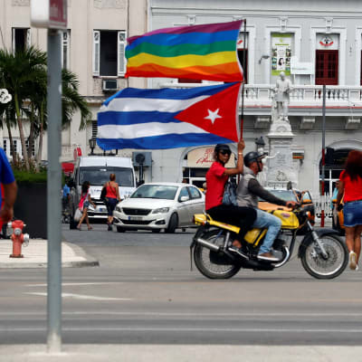 Moottoripyörän kyydissä istuva ihminen heiluttaa Kuubam lippua ja sateenkaarilippua.