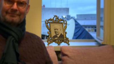 Porträtt på museichef Kai Kartio (suddig) med foto på Amos Andersson i bakgrunden (skarp).