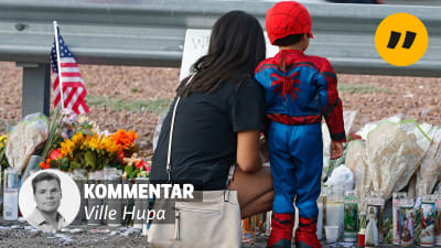 En kvinna och ett barn framför en minnesplats. Det finns mycket ljus och blommor. Text på bilden: Kommentar av Ville Hupa. 