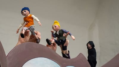 I pjäsen ingår olika dockor. På bilden syns tre dockor, varav de två första klädda i färgglada plagg och den tredje i en mörk kåpa.