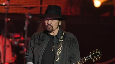 En man med långt svart hår och svart hatt spelar gitarr.