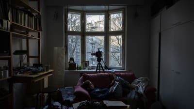 På bilden syns en lägenhet i dunkelt ljus där en person ligger i soffan med datorn i famnen. I fönstret står en kamera riktad ut mot gatan.