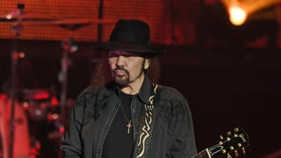 En man med långt svart hår och svart hatt spelar gitarr.