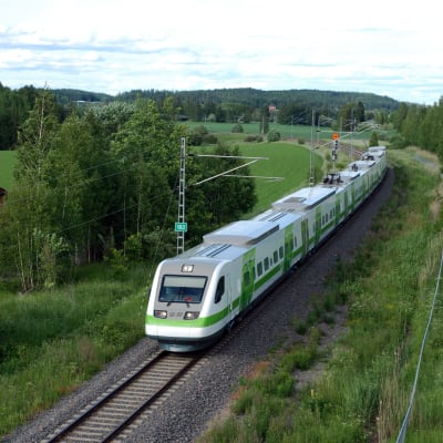 Ett passagerar tåg i vita och gröna färger kör fram i en svag kurva genom ett grönt och lummigt landskap. Järnvägen har bara ett spår.